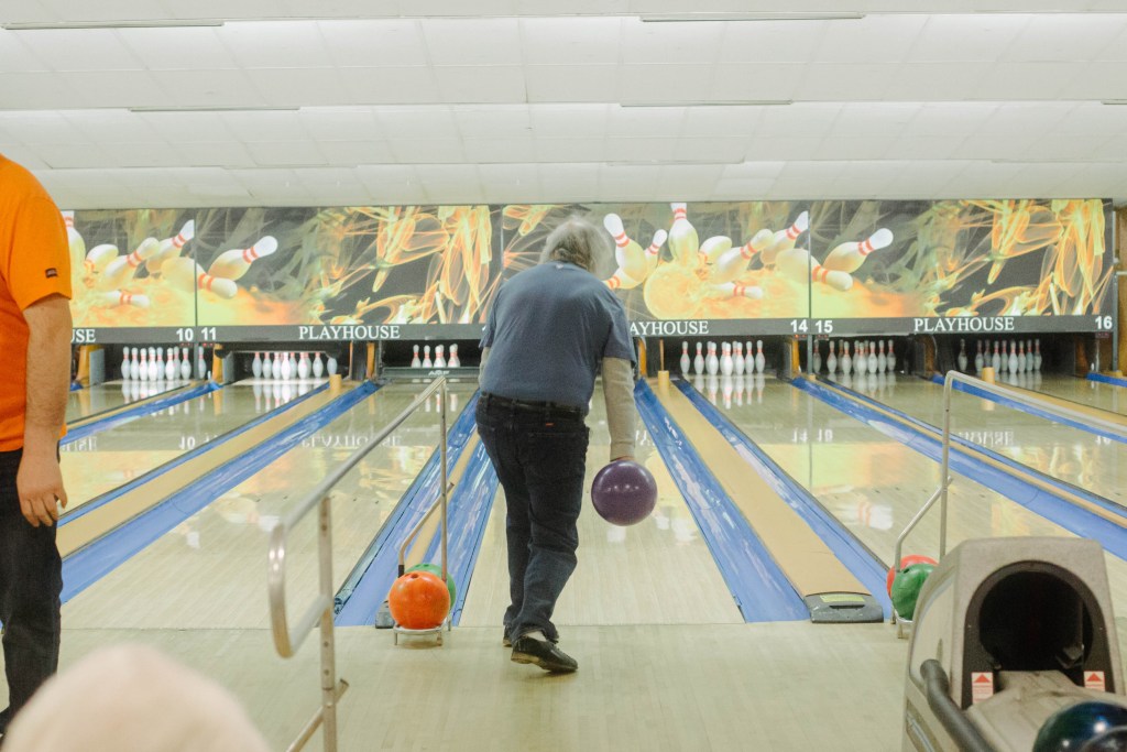 The back of a bowler wearing a gray shirt and black pants at the moment they're about to release a bowling ball down the lane, with the pins at the far end in the background